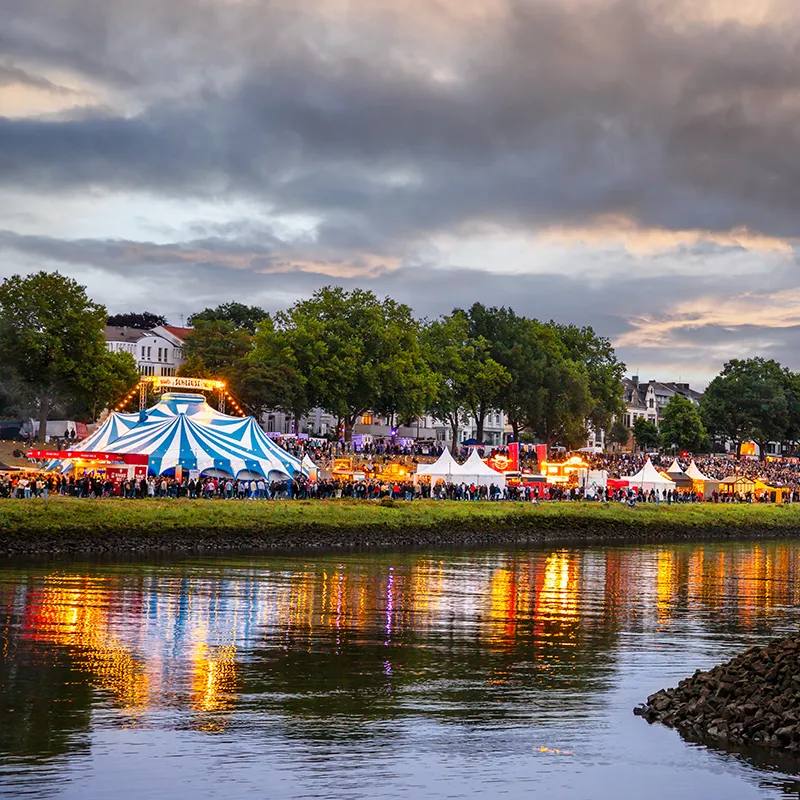 Blick auf die bunten Zelte der stimmungsvoll beleuchteten Breminale am frühen Abend vom gegenüberliegenden Ufer der Weser aus.