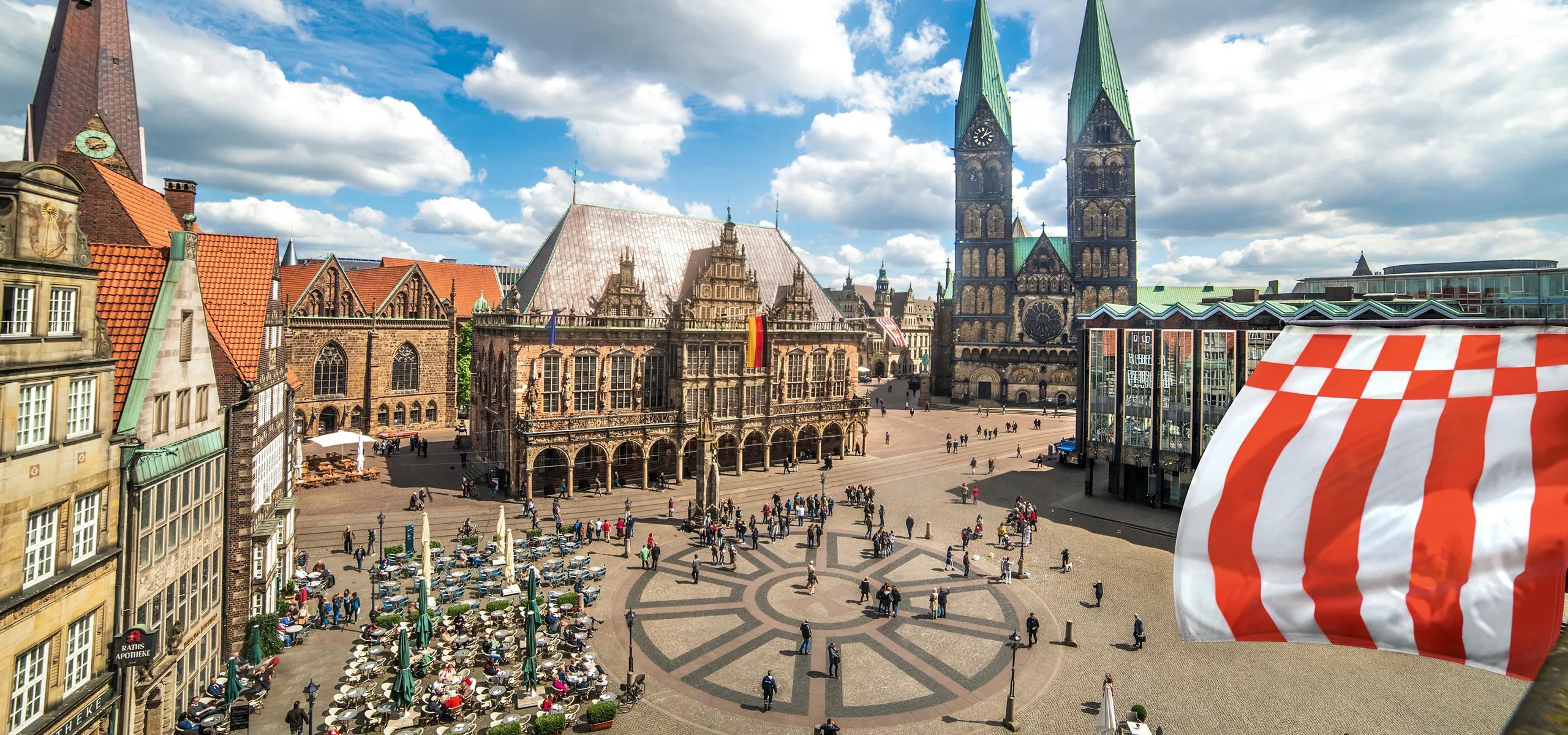 Der Bremer Marktplatz, in der Mitte das beflaggte historische Rathaus, rechts daneben der St. Petri Dom mit zwei Türmen und ganz rechts die Bremische Bürgerschaft.