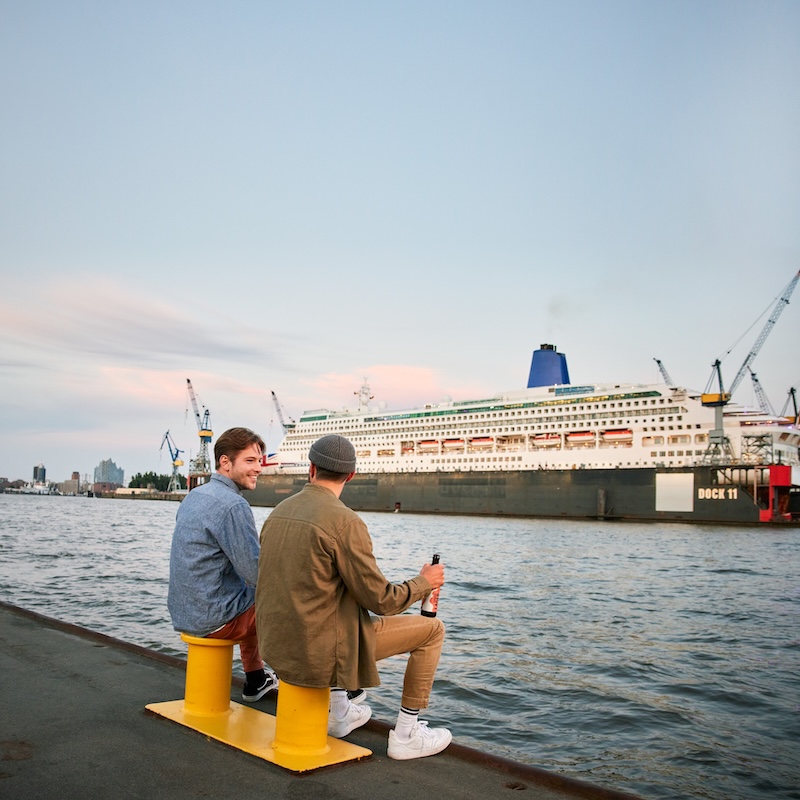 Hamburg versteht sich als 'Das Tor zur Welt', hier dargestellt durch zwei junge Männer, die an einer Kaimauer auf Pollern sitzen und auf ein großes Schiff am gegenüberliegenden Ufer schauen.