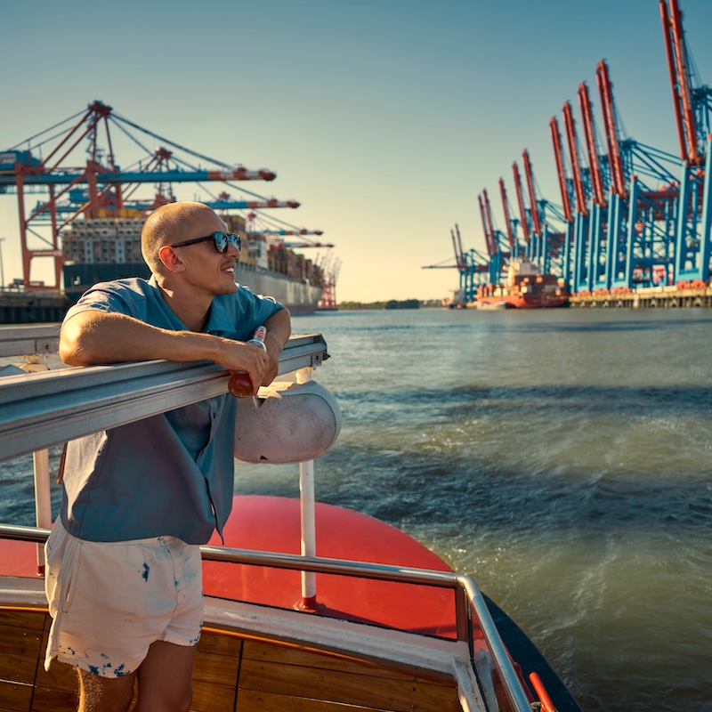 Der Hamburger Hafen vom Wasser aus. Im Hintergrund zahlreiche Container-Verladekräne und ein Containerschiff am Kai.