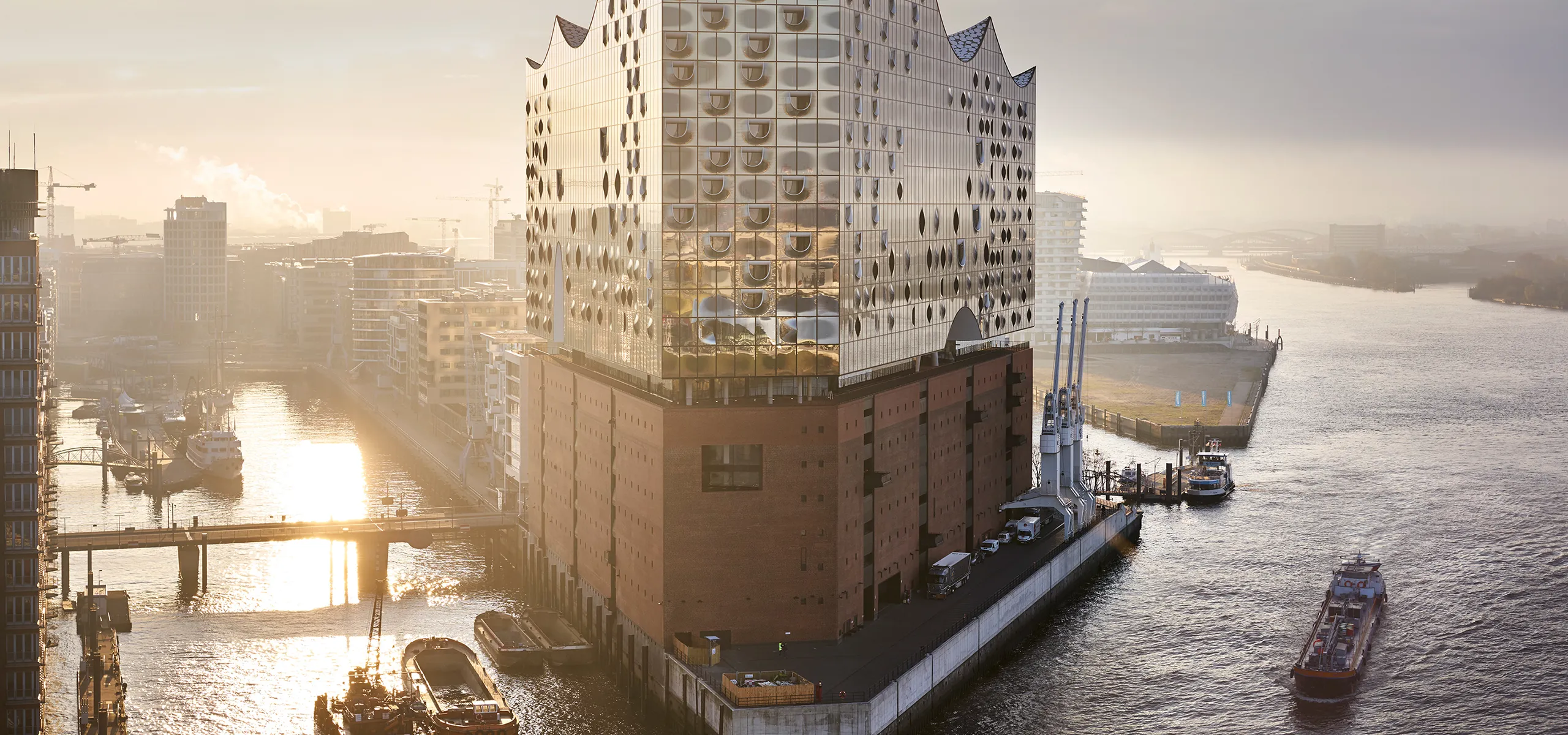 Ein imposantes Gebäude direkt an einem großen Fluss, unten aus Backstein, oben ab der Mitte aus einer Glasfront mit geschwungenen Öffnungen.