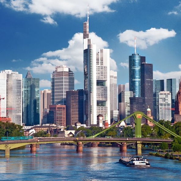 Die Skyline von Frankfurt am Main mit zahlreichen Hochhäusern, deren Glasfassaden und Fenster im Sonnenlicht leuchten und dem Fluss 'Main' im Vordergrund, symbolisieren die Position der Stadt als führendes und global vernetztes Finanzzentrum Europas.