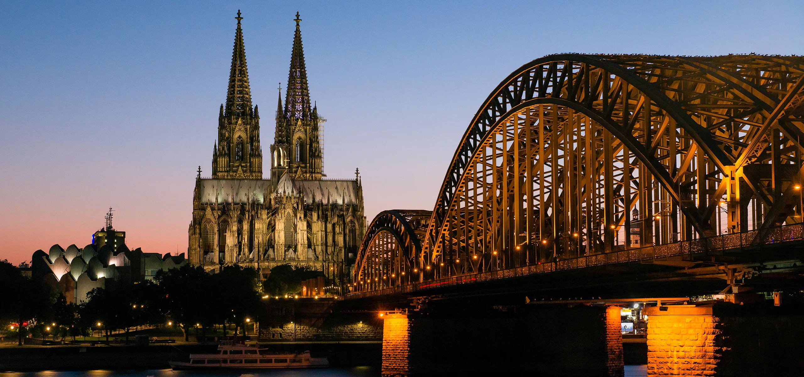 Der erleuchtete Kölner Dom bei Nacht, rechts eine Bogenbrücke.