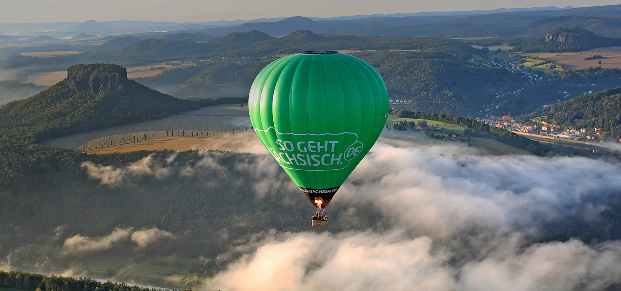 Ein grüner Heißluftballon über leicht hügeligen Wiesen und Feldern, darunter ein paar fluffige Wolken.