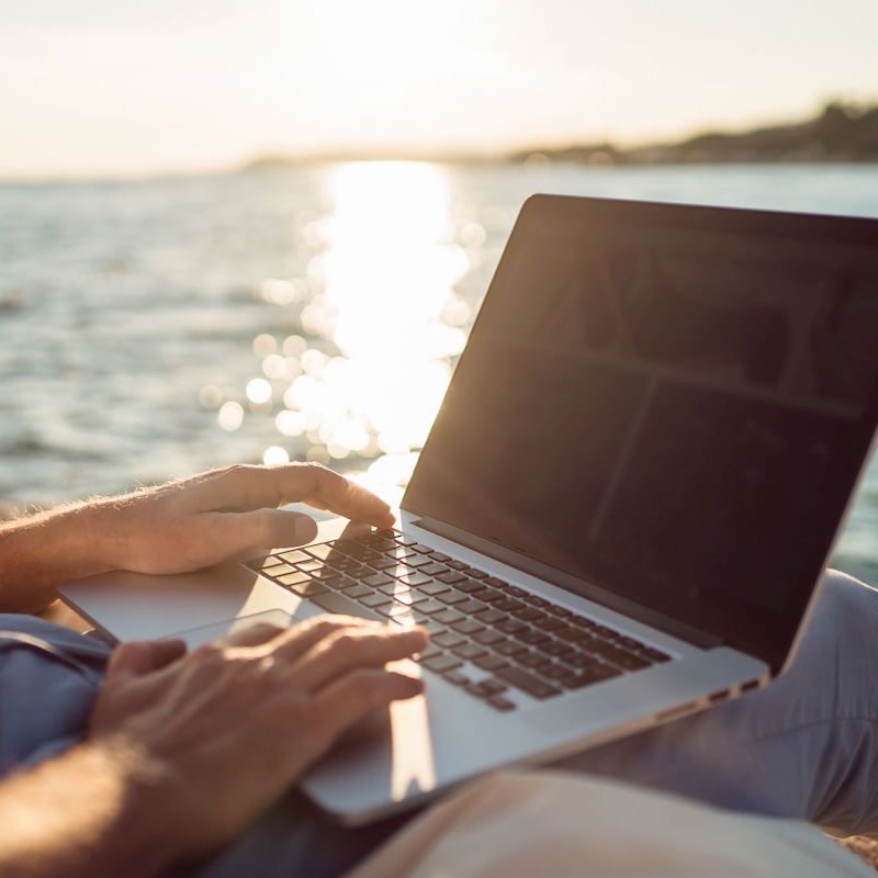 Eine Person arbeitet in der Abendsonne mit einem mobilen Computer an einem See.