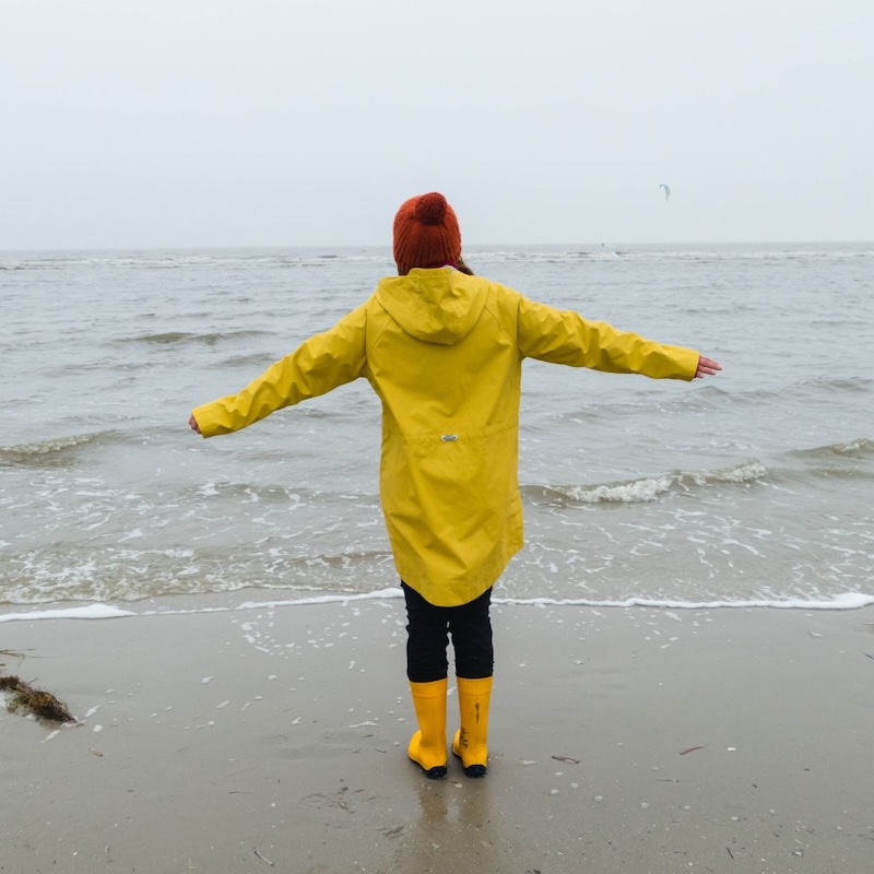 Eine Person in einem gelben Regenmantel steht bei schlechtem Wetter an einem Strand und genießt die Situation mit offenen Armen.