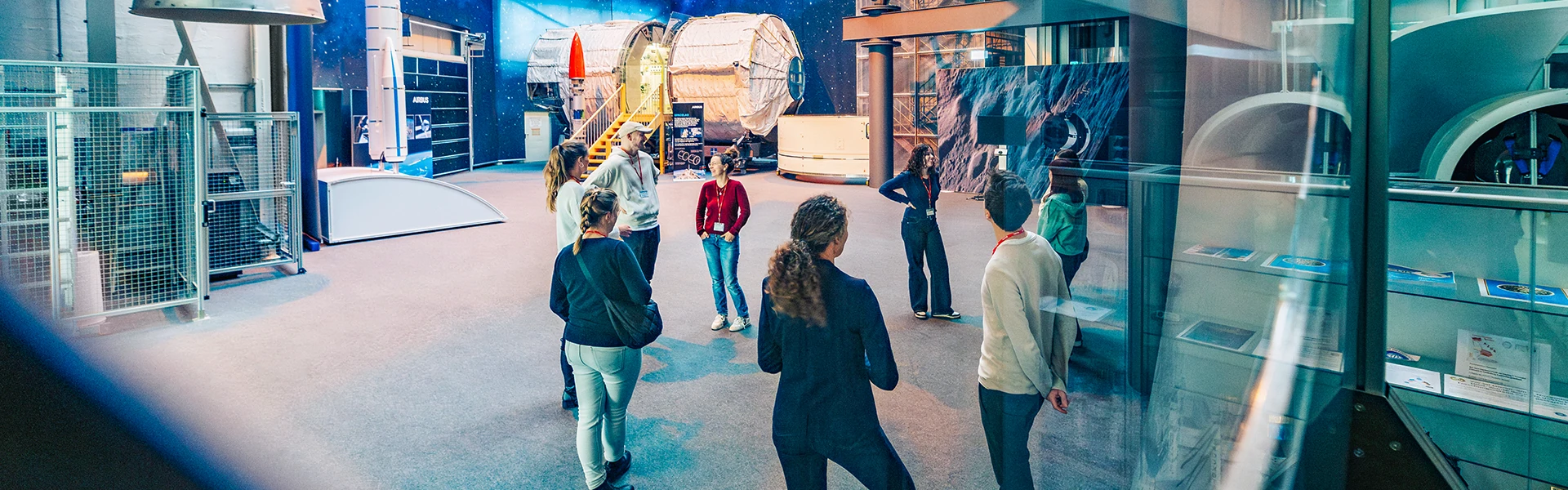 Menschen stehen bei einer Führung durch ein Weltraumsetting im Kreis, unterhalten sich und betrachten die Exponate. Im Hintergrund ein Raketenmodell.