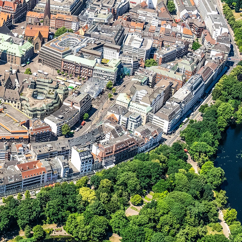 Ein Luftbild mit einer Straße in der Mitte, die einen bebauten Bereich oben von einem Grünzug unten trennt.