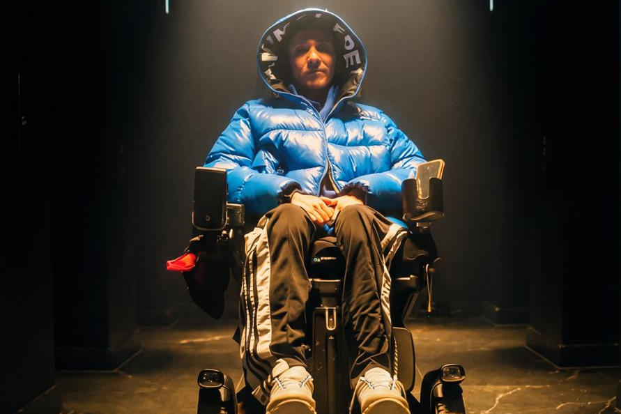 Ein stylisch gekleideter junger Mann im Licht eines Scheinwerfers. Er sitzt im Rollstuhl.