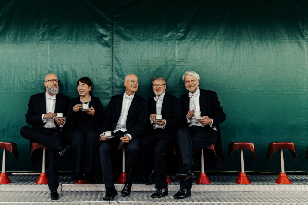 Mitglieder eines Orchesters trinken vor einer grünen Wand einen Kaffee.