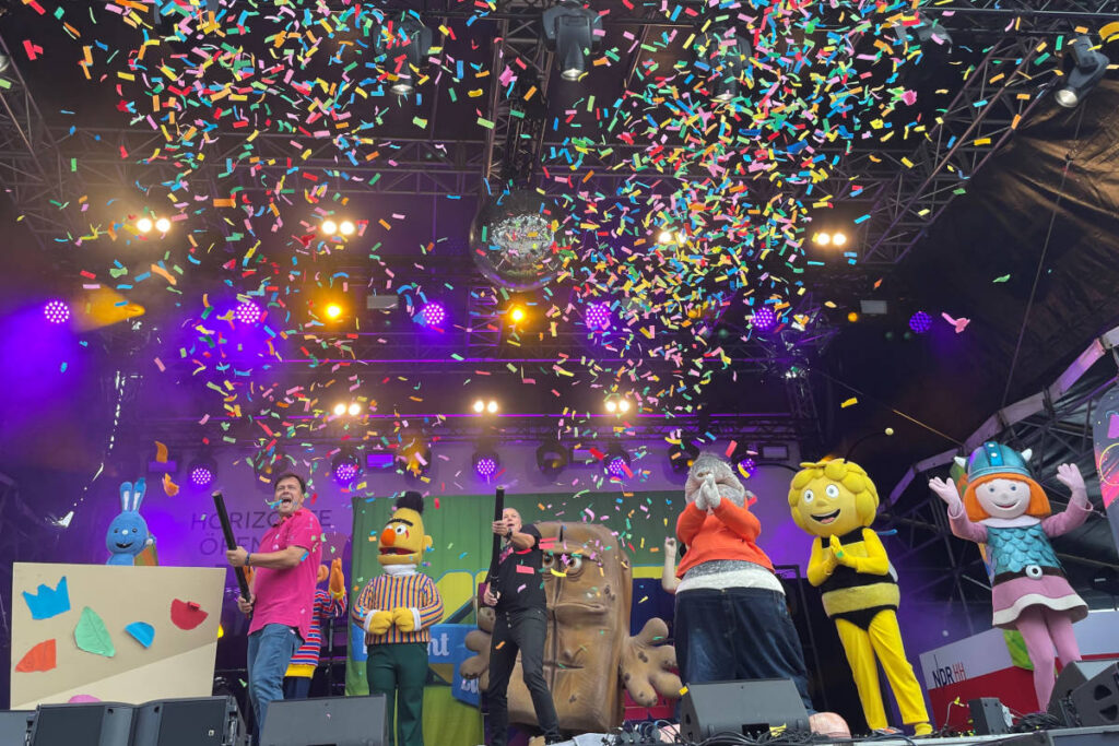 Verschiedene Figuren aus dem Kinderprogramm stehen auf einer Bühne und begeistern das bunt gemischte Publikum.
