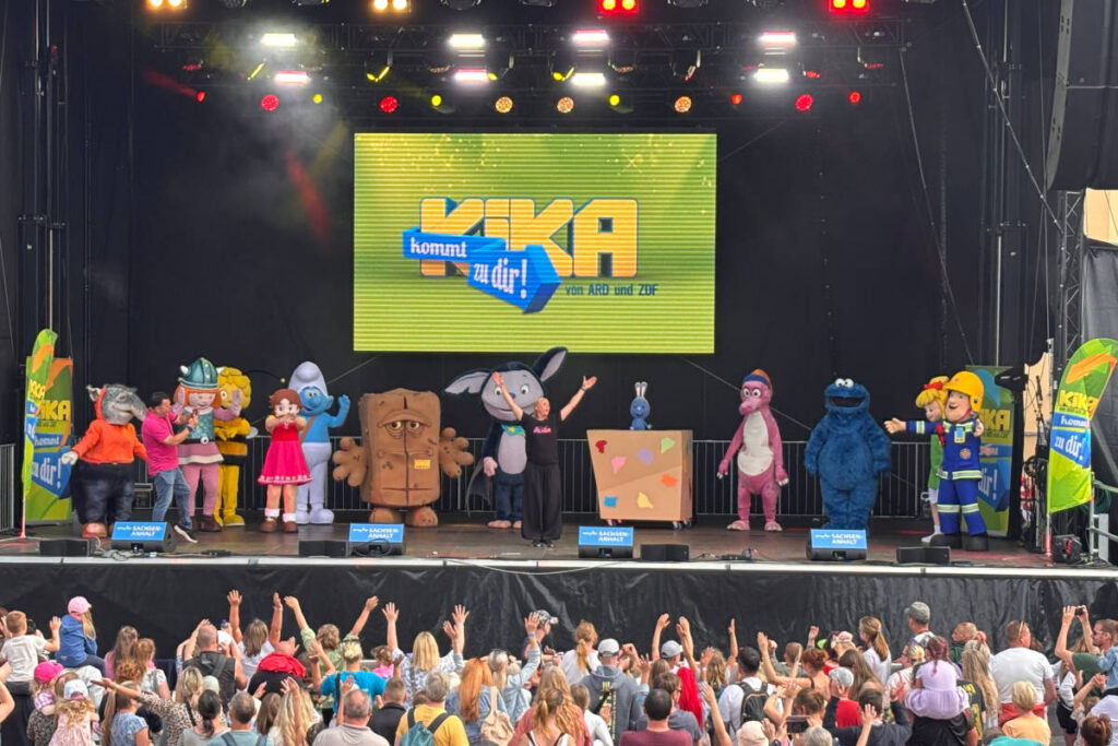Verschiedene Figuren aus dem Kinderprogramm stehen auf einer Bühne und begeistern das bunt gemischte Publikum.