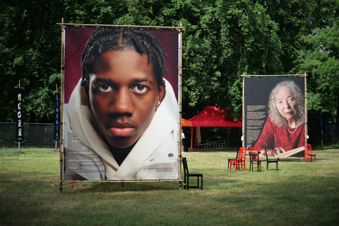 Große Plakate mit Portraitaufnahmen und Geschichten auf einer Rasenfläche. Stühle laden zum Verweilen ein.