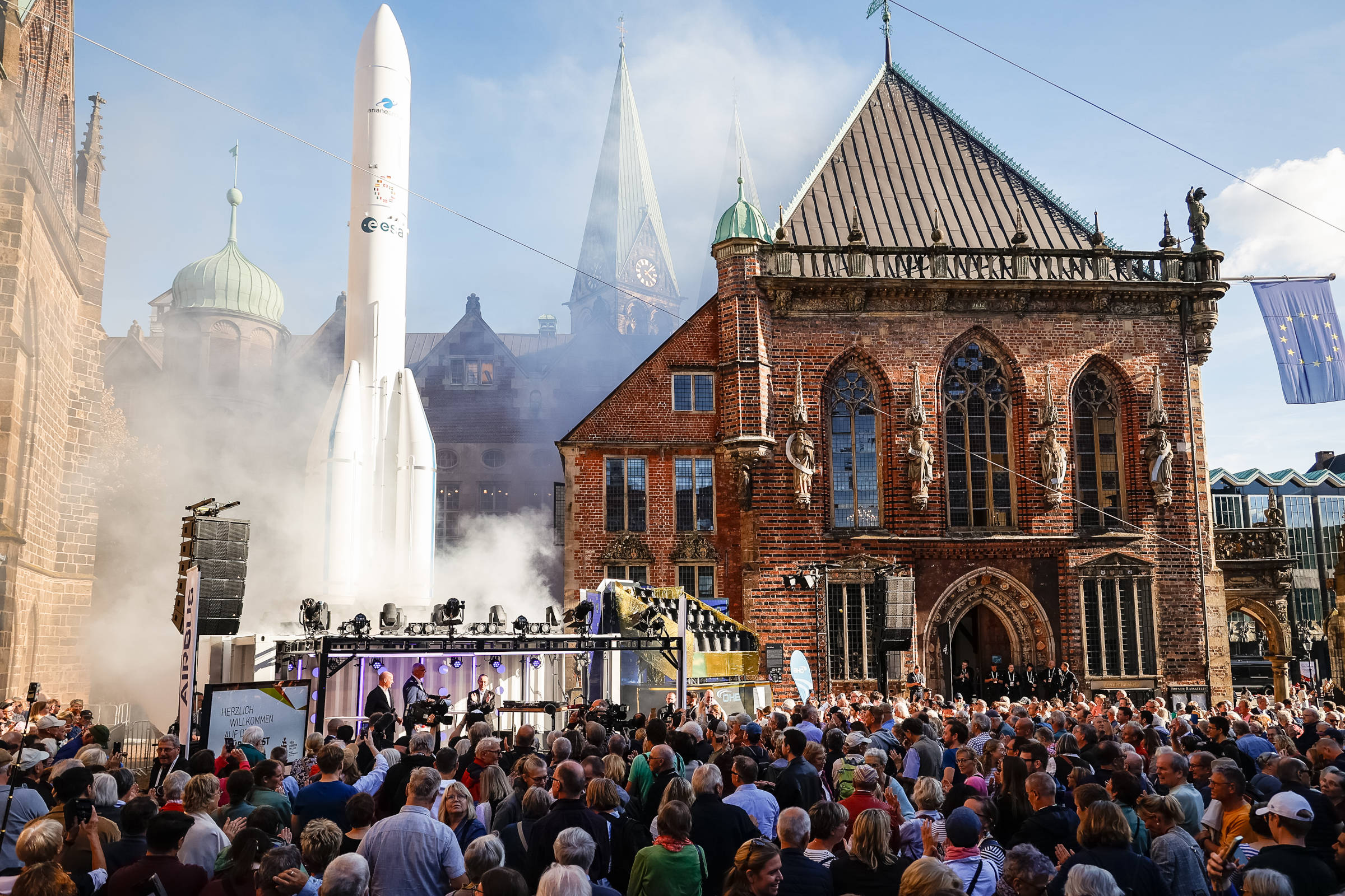 Eine Menschenmenge vor einer Bühne, im Hintergrund ein Modell einer Ariane-Rakete und das Bremer Rathaus.